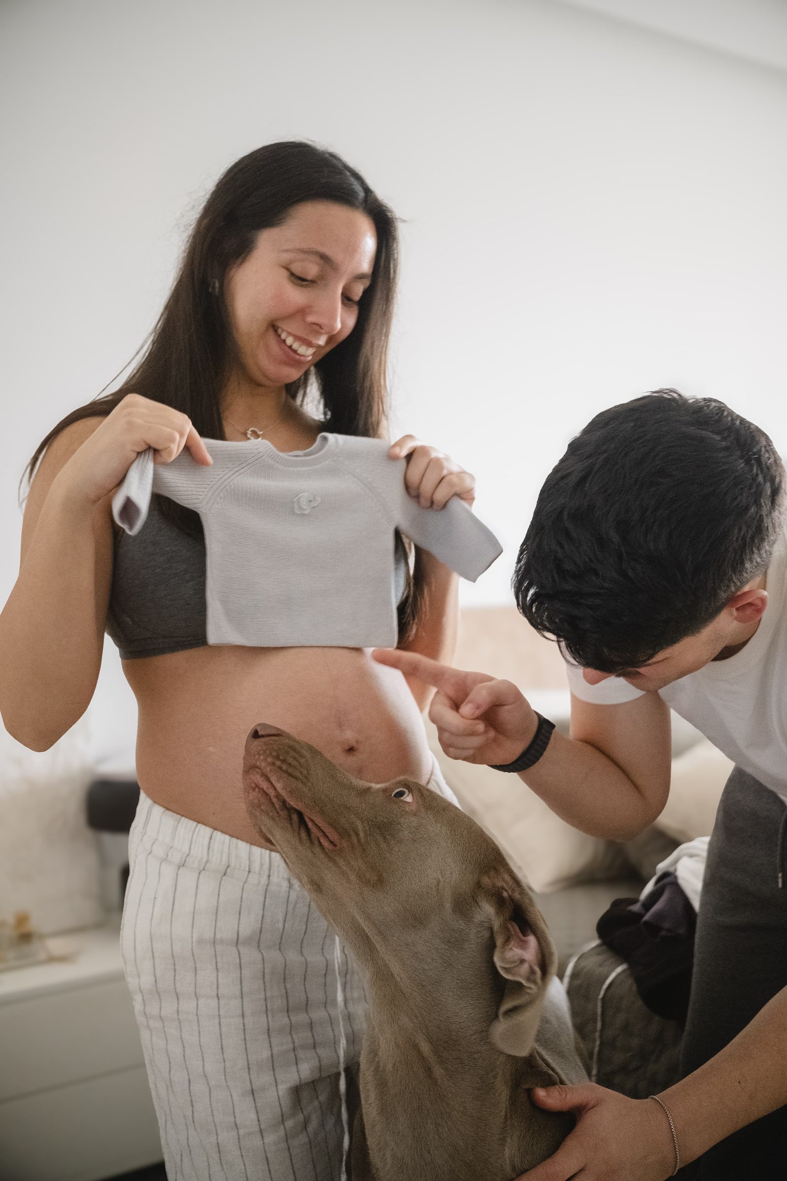 Casal à espera de um bebé ri-se enquanto mostra um pequeno casaco de bebé, com o cão a olhar para eles